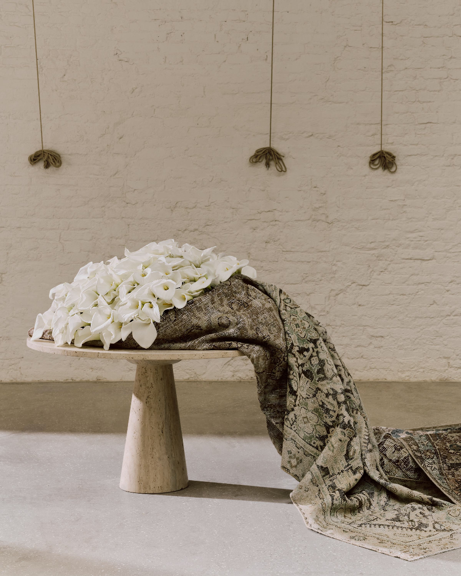 White flowers on a stone pedestal with a patterned fabric draped over it against a beige brick wall.
